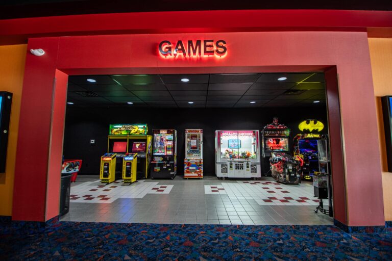 An image of an arcade entrance with a red sign above reading 'GAMES'. Inside are various arcade machines, including claw machines and video games, set on a tiled floor with colorful carpet leading into the area.