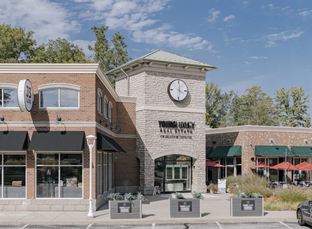 A brick building with a prominent clock tower and signage for Young Luxury Real Estate. The building features large windows, black awnings, and outdoor planters with greenery. Adjacent to it is an area with red umbrellas, indicating a possible outdoor dining space. The sky is clear with scattered clouds and trees are visible in the background.