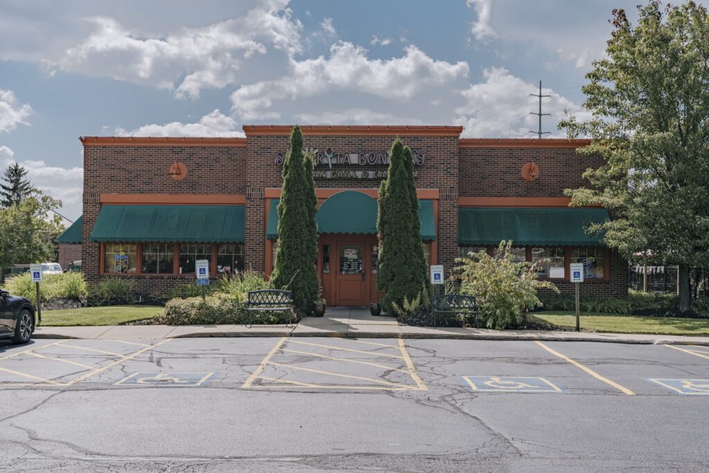 The image shows the exterior of a brick restaurant with a green awning over the windows and entrance. There are two tall bushes flanking the entrance and handicap parking spaces marked in front of the building. The sky is partly cloudy and there is a tree on the right side of the image.