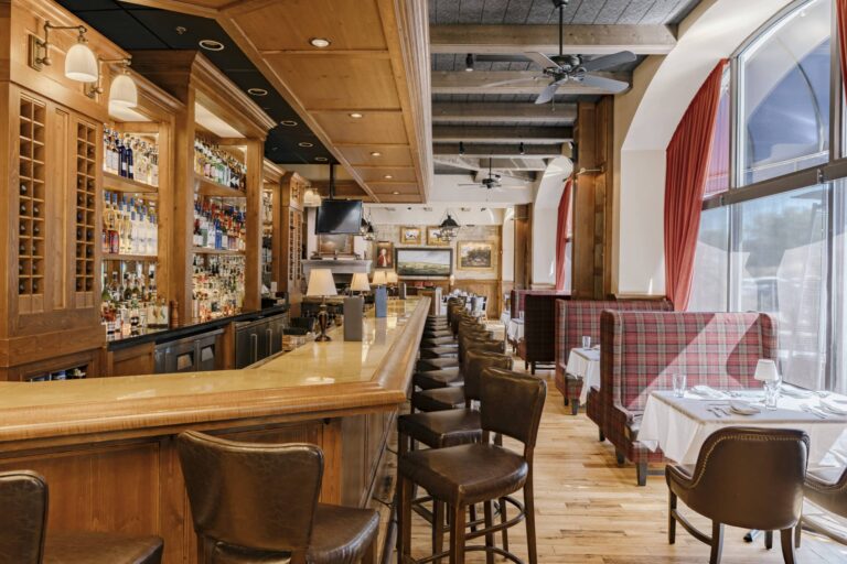 A cozy restaurant bar interior featuring a long wooden counter with leather bar stools arranged neatly. Behind the bar, shelves are stocked with various liquor bottles. To the right, there are wooden booths with checkered upholstery and tables set with white tablecloths, glasses, and cutlery. Large windows with red curtains allow natural light to stream in, highlighting the warm wooden floors and ceiling fans overhead.