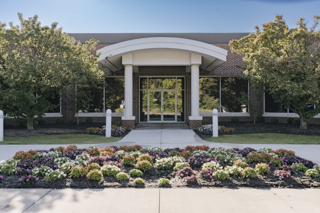 A modern building entrance featuring a grand entrance with white pillars and large glass doors. The foreground displays a well-maintained landscaped garden with vibrant flowers in various colors, bordered by neatly trimmed grass and flanked by two trees.