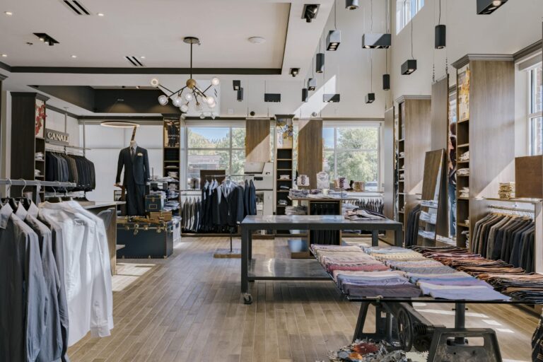 Interior of a modern menswear boutique featuring neatly arranged racks of suits, dress shirts, and ties. A central table displays colorful fabrics, while a mannequin showcases a formal suit. The store has large windows allowing natural light inside and a stylish chandelier hanging from the ceiling.