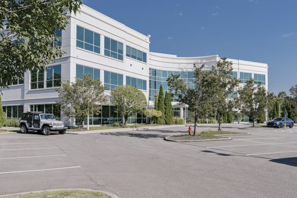 A modern office building with large glass windows and a white facade. The building is surrounded by trees and landscaped bushes. In the foreground, there is a parking lot with two vehicles parked, a grey SUV and a dark sedan. A blue sky is visible in the background.