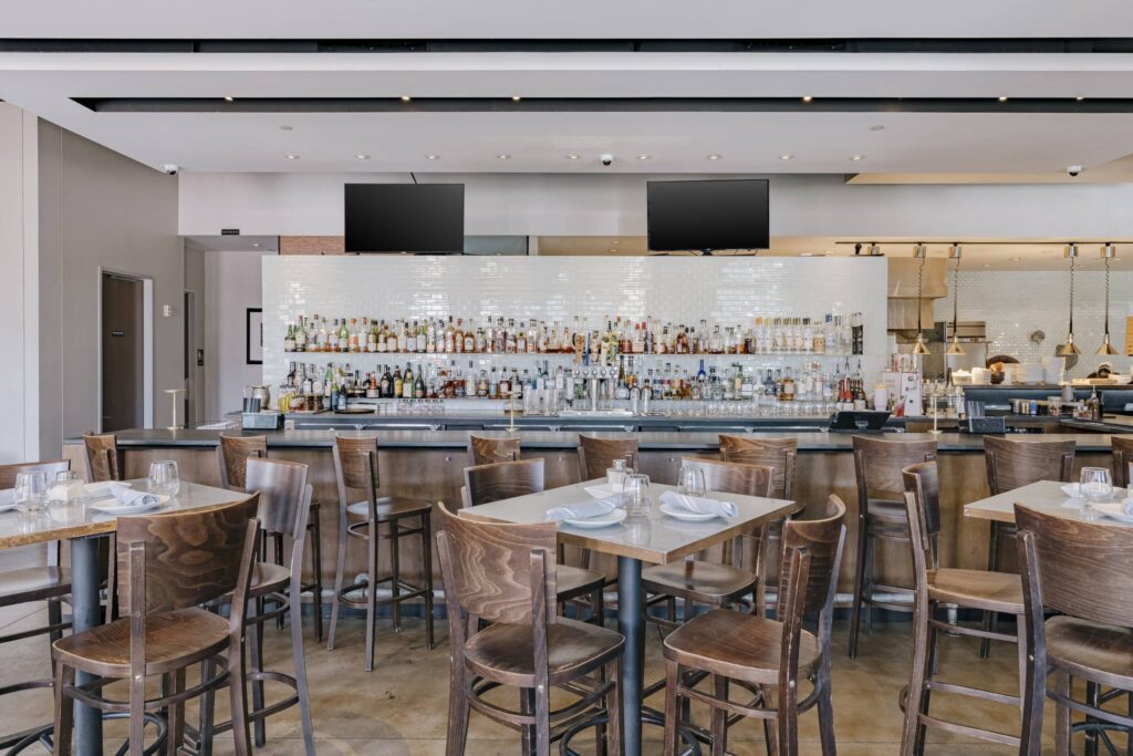 A modern restaurant interior featuring a well-stocked bar with rows of various liquor bottles on display. Wooden tables and chairs are set with napkins and glasses in the foreground. The overall ambiance is bright and inviting, with two large screens mounted above the bar.