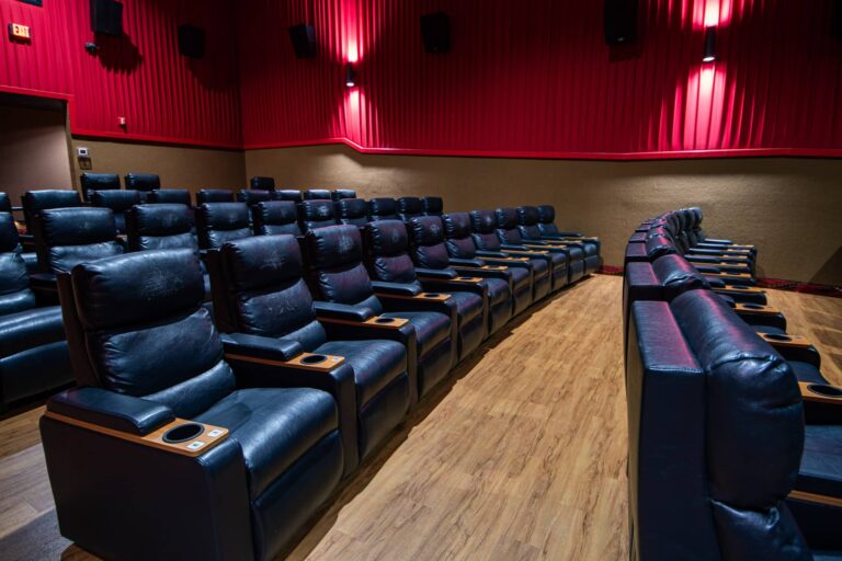 Interior of a modern movie theater featuring rows of comfortable black leather reclining seats with wooden armrests, cup holders, and a warm-toned wooden floor. The theater walls are adorned with red acoustic panels, and a partially visible exit sign is on the left side.