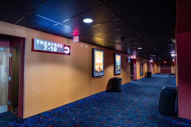 A dimly lit movie theater hallway featuring a sign indicating 'Theatres 7-10' with an arrow pointing right. The walls are adorned with movie posters, and there is an exit sign above a closed door. The floor is covered with a patterned carpet, and trash cans line the hallway.