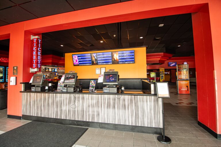 Interior view of a movie theater ticket counter with multiple electronic terminals and neon 'Tickets' signs. The counter is framed by a red and orange color scheme, and overhead displays show current movie schedules.