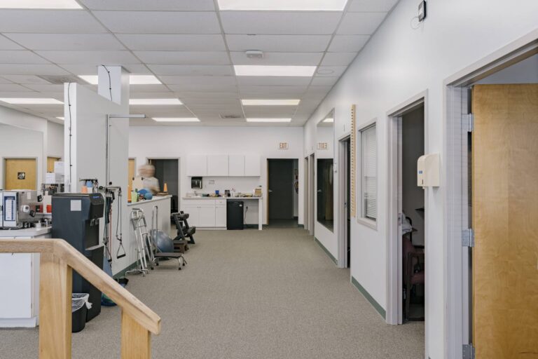 Interior view of a physical therapy clinic featuring equipment such as a treadmill, balance balls, and exercise tools along a hallway. The space has a neutral color scheme with white walls and grey carpet, with several rooms lining the corridor.