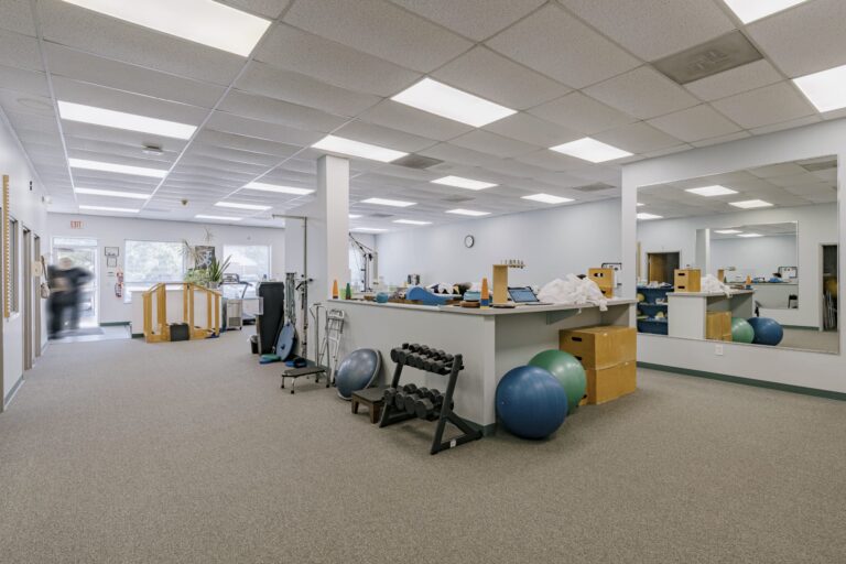Interior of a physical therapy gym featuring exercise equipment like dumbbells, yoga balls, and physical therapy tools. The room has a carpeted floor, mirrors on the walls, and fluorescent lighting. A person is seen walking towards an exit in the background, and windows allow natural light to enter.