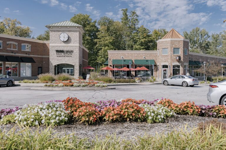 The image depicts the exterior of a shopping center with a clock tower and brick buildings. Colorful flowers are landscaped in the foreground, and there are parked cars and outdoor seating with umbrellas at the complex.